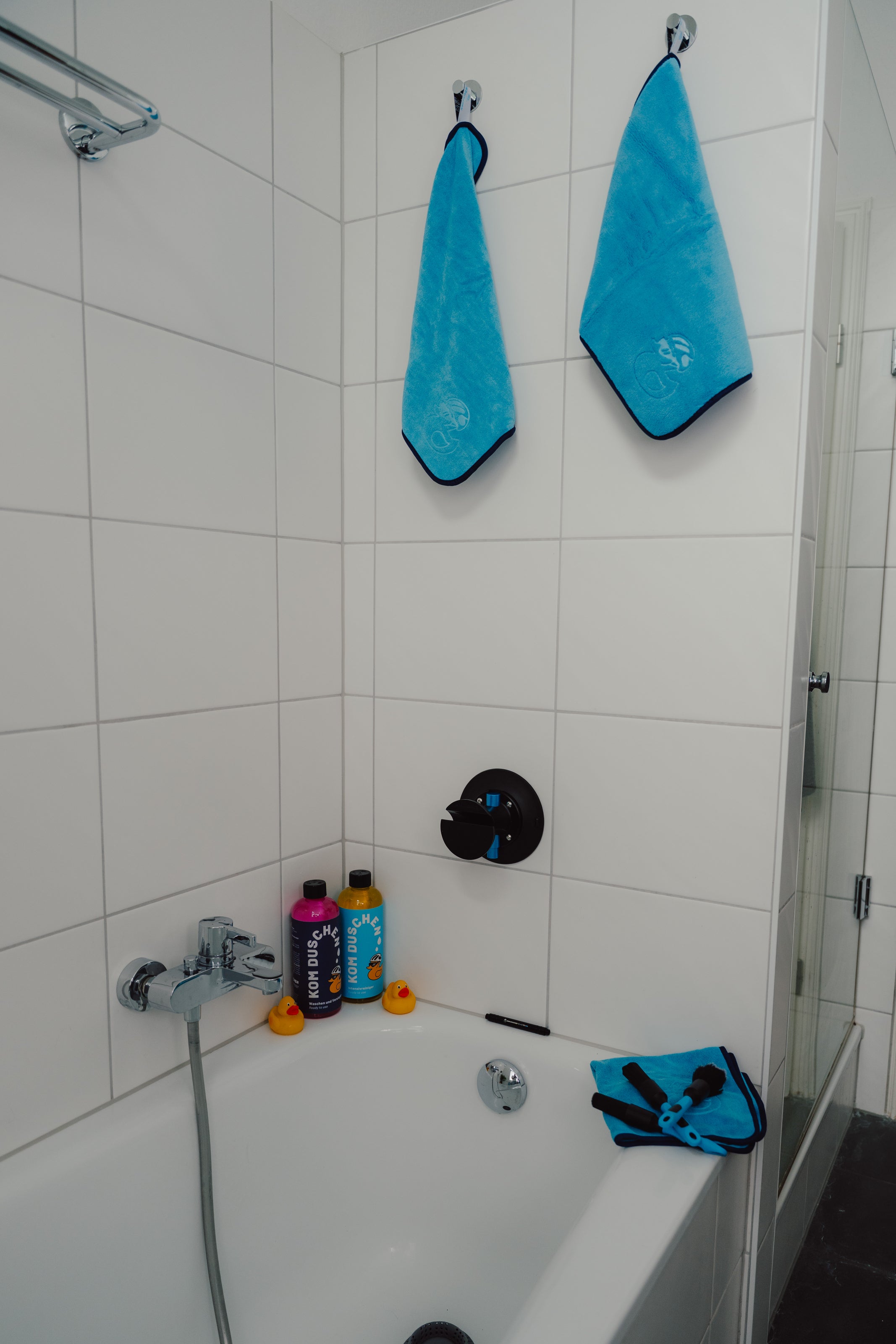Ein weiß gefliestes Badezimmer mit einer Badewanne, drei Flaschen und zwei Gummienten am Wannenrand sowie mehreren blauen Handtüchern der Marke „Mikrofasertuch – Reinigungstücher 400 g/m² (4 Stück)“, die an Haken hängen und über den Wannenrand drapiert sind.
