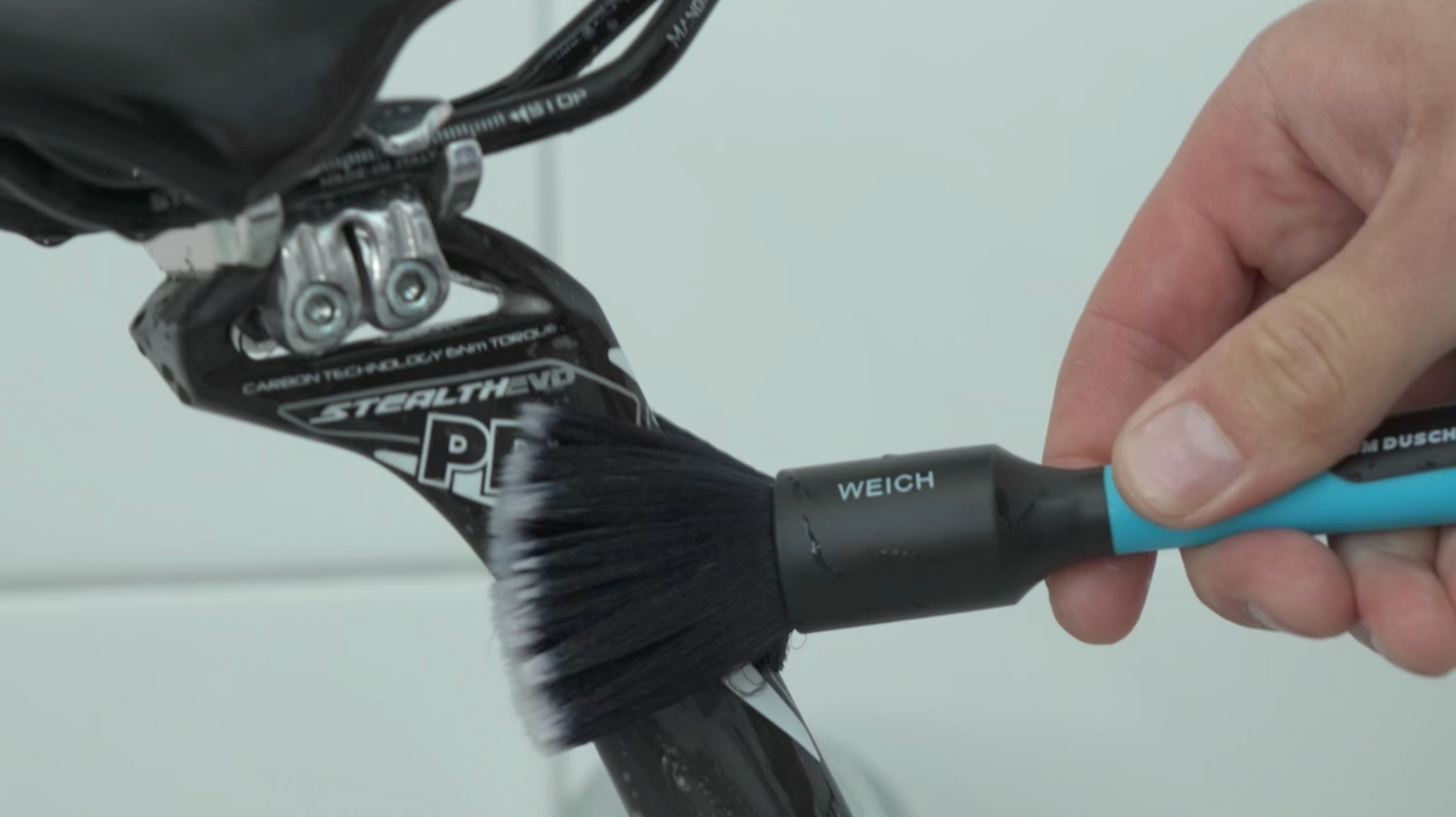 Eine Hand benutzt eine Bürste aus dem Pinselset - Fahrrad Detailing, um den Rahmen eines schwarzen Fahrrads in der Nähe der Sattelstütze zu reinigen.