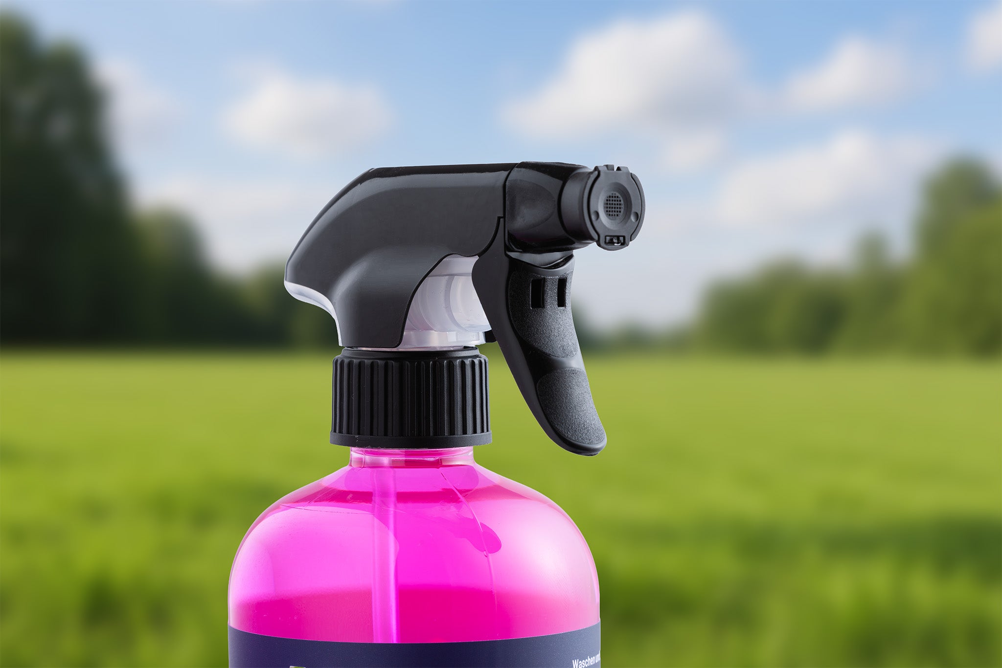 Eine Nahaufnahme einer Sprühflasche mit schwarzer Düse und leuchtend rosa "Waschen und Versiegeln - Ready to Use" im Inneren vor einem unscharfen Hintergrund aus Gras und Bäumen unter einem teilweise bewölkten Himmel.