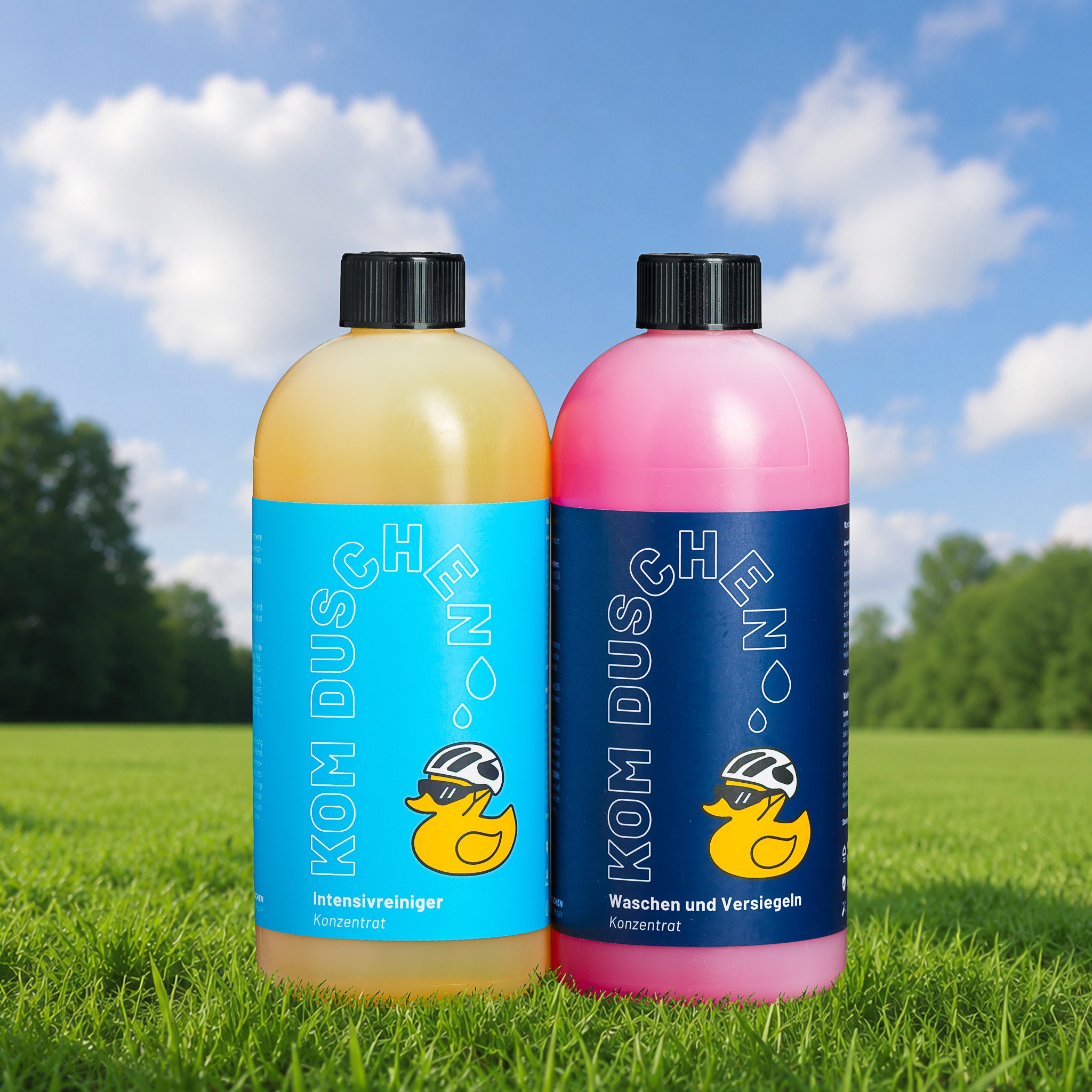 Zwei Flaschen aus dem „Essential Konzentrate“-Set mit Entenmotiven stehen auf Gras, im Hintergrund sind grüne Bäume und ein blauer Himmel mit Wolken zu sehen. Die Flasche auf der linken Seite ist gelb, die auf der rechten Seite rosa.