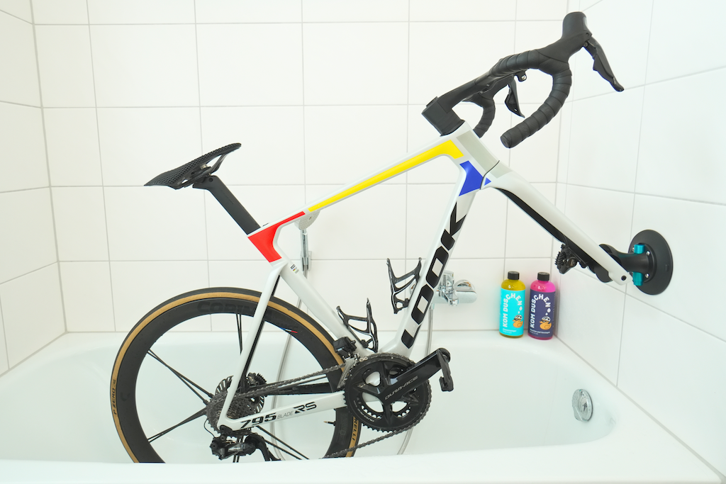 Ein Saugnapfhalter Fahrrad ist in einer weißen Badewanne in einem gefliesten Badezimmer zum einfachen Rennrad waschen angebracht. Zwei bunte Flaschen sind auf dem Wannenrand in der Nähe des Hinterrades platziert.