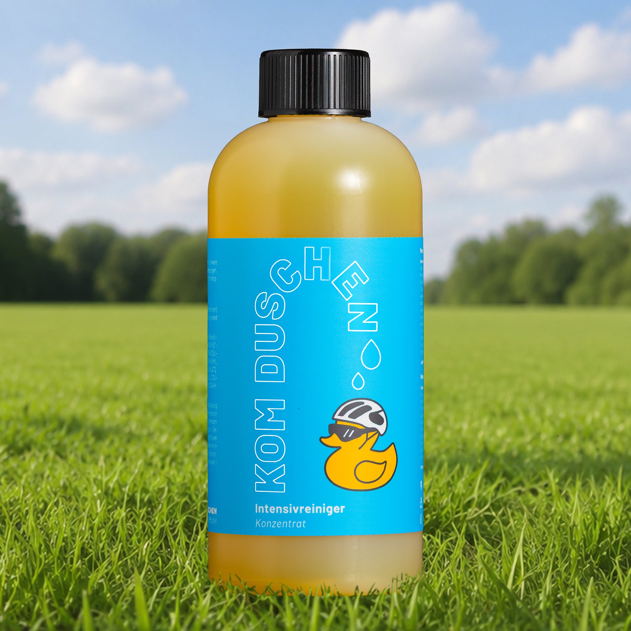 Eine Flasche mit der Aufschrift „Intensivreiniger – Konzentrat“, die ein blaues Etikett, eine Entenillustration und einen schwarzen Verschluss aufweist, steht auf grünem Gras vor einem unscharfen Hintergrund mit Landschaft und Himmel – ideal für alle, die ihr Fahrrad nach einer langen Tour reinigen möchten.