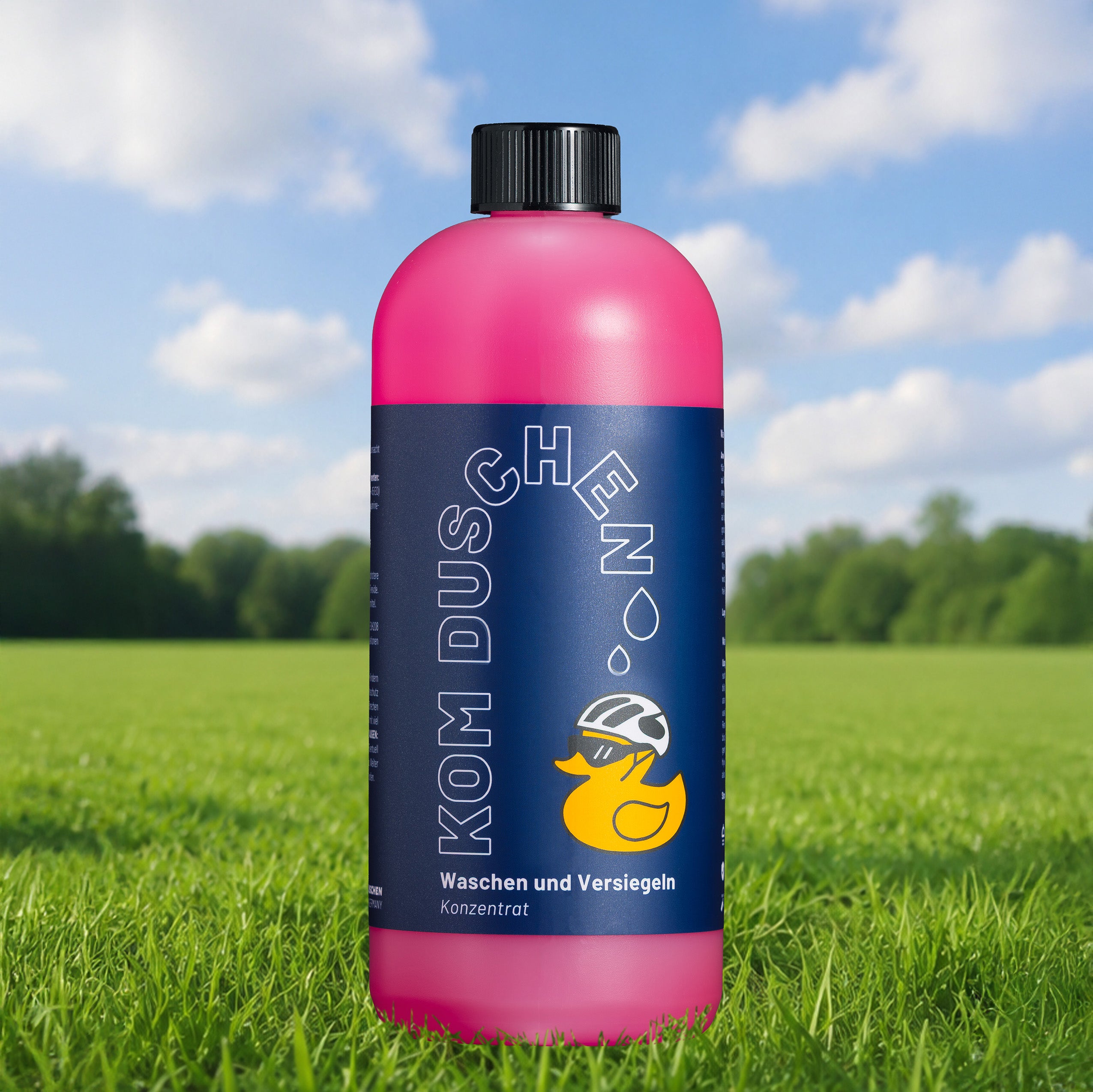 Eine rosa Flasche mit einem blauen Etikett, auf dem „Kom Duschen“ steht und eine gelbe Ente abgebildet ist, vor einem Hintergrund aus Gras, Bäumen und blauem Himmel – dieses Produkt – „Waschen und Versiegeln – Konzentrat“ – eignet sich perfekt für die Fahrradwäsche und bietet bei jeder Wäsche sanften Lackschutz.