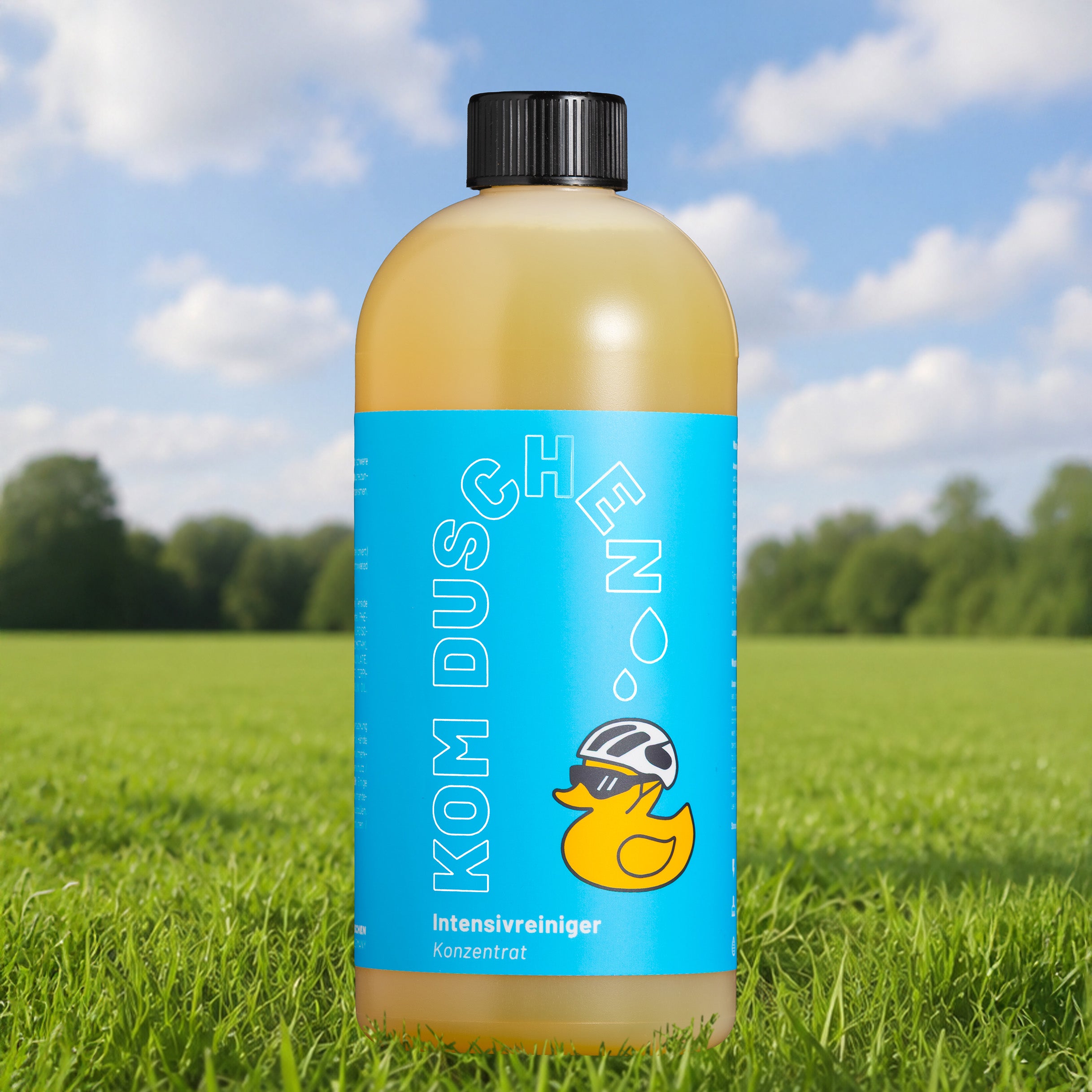 Auf grünem Gras, hinter dem Bäume und ein blauer Himmel zu sehen sind, steht eine Flasche mit einem blauen Etikett, auf dem eine Comic-Ente mit Helm abgebildet ist. Auf dem Etikett steht „KOM DUSCHEN“ und „Intensivreiniger – Konzentrat“ – damit ist es der perfekte Intensivreiniger – Konzentrat für Ihre nächste Fahrradwäsche.
